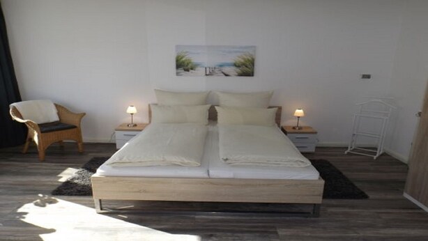 A double bed with two nightstands, a chair, a small table, and a painting on the wall. - Bright and modern apartment right on the North Sea (Nordstrand)
