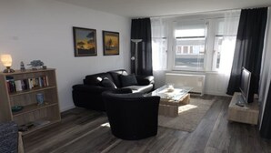 A modern living room with a black sofa, a glass coffee table, and a bookshelf. - Bright and modern apartment right on the North Sea (Nordstrand)