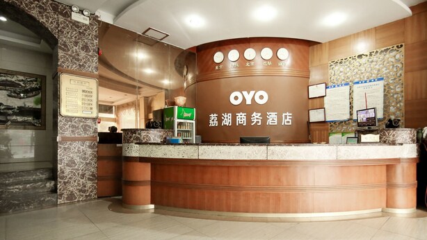 A reception desk with a wooden finish and a sign that reads "OYO". There are several clocks on the wall above the desk, and a bulletin board with papers and a small screen. - OYO 8027 Li Hu Business Hotel (Shenzhen)