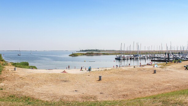 En strand med en småbåtshamn, en sandstrand och vindkraftverk i fjärran.
