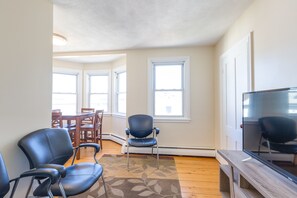 A living room with a large flat-screen TV, a wooden coffee table, and a patterned rug. There are two black chairs and a dining table with chairs in the background. - Chestnut Hill Apts by Starlight Suites (Brookline)
