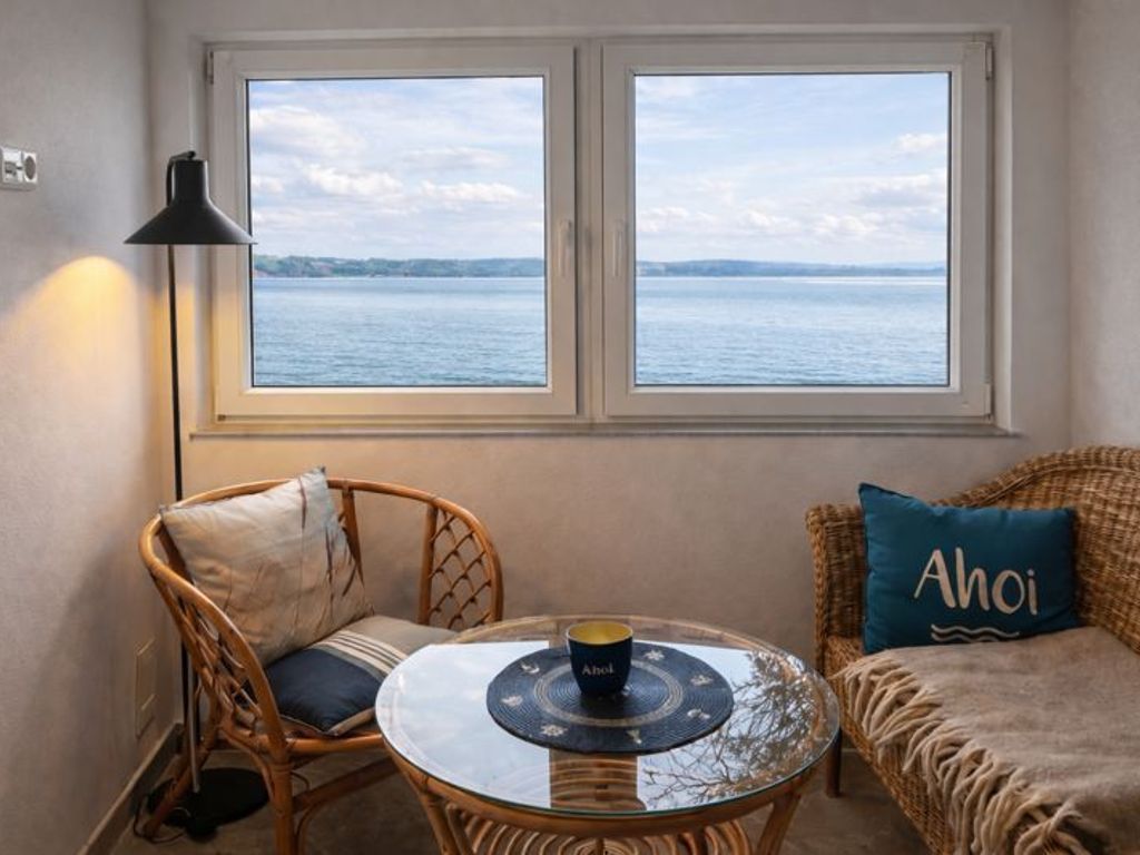Ein Zimmer mit zwei Fenstern und Blick auf ein Gewässer, ein Korbstuhl, ein runder Glastisch mit Untersetzer und eine Couch mit einem Kissen mit der Aufschrift 'Ahoi'.