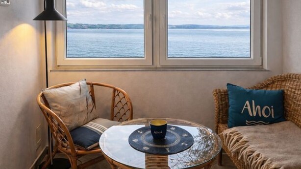 A room with two windows overlooking a body of water, a wicker chair, a round glass table with a coaster, and a sofa with a cushion labeled "Ahoi". - Apartment 2 - Ahoi-Marie (Middelhagen)