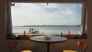 A room with a large window overlooking a lake, a table with chairs, and decorative items on the windowsill. - Apartment 2 - Ahoi-Marie (Middelhagen)