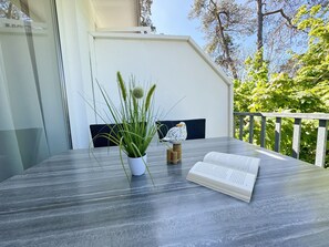Une table ornée d’une plante en pot et d’une statuette d’oiseau, avec un livre ouvert, et une vue sur des arbres et une rambarde blanche.
