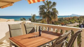 A balcony with a wooden table and chairs, overlooking a coastal view with palm trees and a mountain in the distance. - 2 bedroom lovely apartment in Piles (Piles)