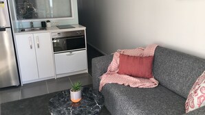 A compact kitchen with white cabinets, a built-in oven, and a window above the sink. A gray sofa with a pink throw blanket and pillows is positioned against the wall. - Altitude (Auckland)