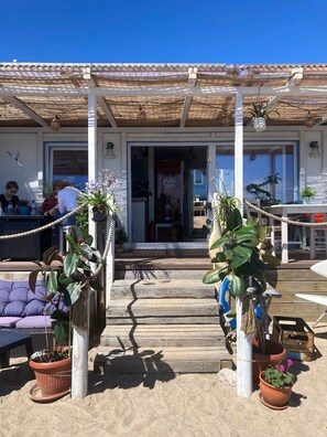 A covered outdoor seating area with potted plants, a wooden deck, and a building entrance.