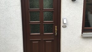 Eine braune Tür mit Fensterglas, Klingel und Wandleuchte.