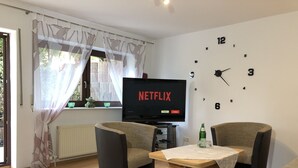 Ein Wohnzimmer mit Fernseher, auf dem 'NETFLIX' angezeigt wird, zwei Sesseln, einem kleinen Tisch und einer Wanduhr.