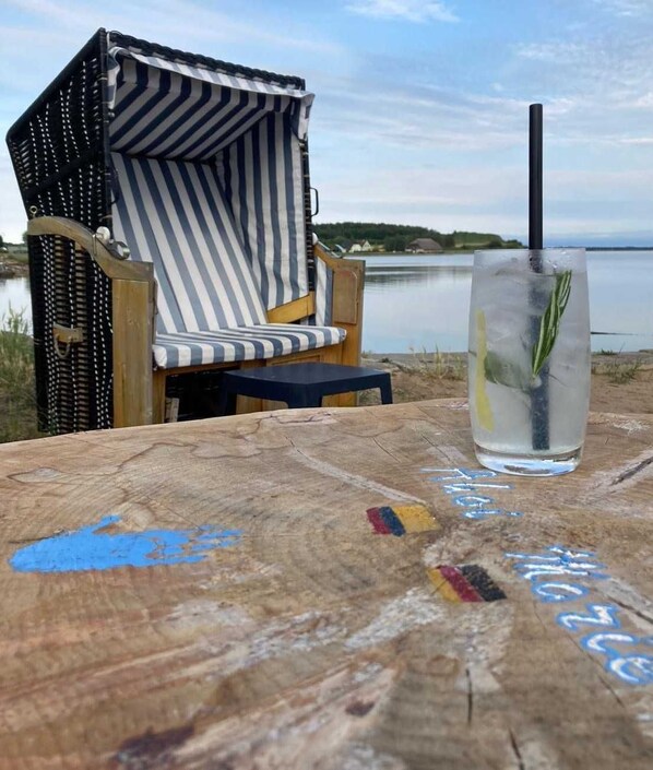Une chaise de plage à rayures, un verre d’eau avec une paille et une branche de romarin, et une table en bois ornée de motifs peints.