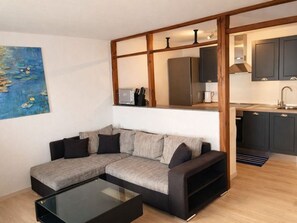 Un salon moderne avec un canapé d’angle, une table basse en verre et un coin cuisine visible à travers une cloison en bois.