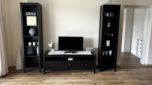 A living room with a TV on a black TV stand, two black bookshelves with decorative items, and a wooden floor. - Sunny 2 room apartment with a fantastic view of Lake Millstatt (Millstatt)