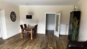 A living room with a wooden dining table, chairs, a coffee table, a couch, a wall clock, and a cabinet with plants. - Sunny 2 room apartment with a fantastic view of Lake Millstatt (Millstatt)
