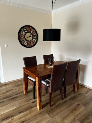 A dining area with a wooden table and wicker chairs, a wall clock, and a black pendant light.