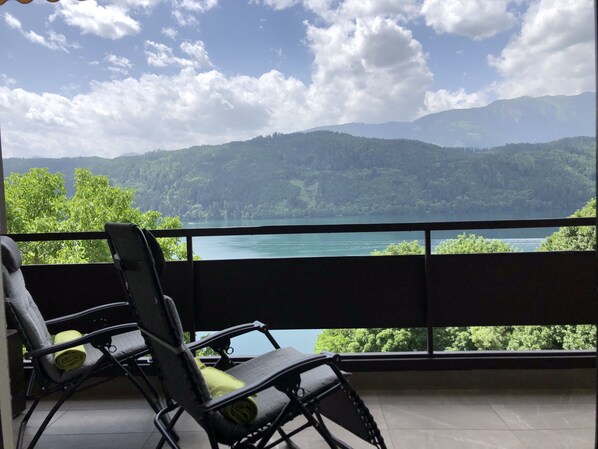 Un balcón con dos sillas y una mesa, con vista a un lago y colinas boscosas.