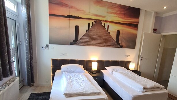 A hotel room with two beds, a large sunset painting on the wall, and a view of the sea. - Apartment-32609de (Hüllhorst)