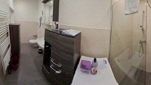 A bathroom with a toilet, sink, mirror, and shower. There is a red bucket and a bottle of hand soap on the counter. - Apartment-32609de (Hüllhorst)