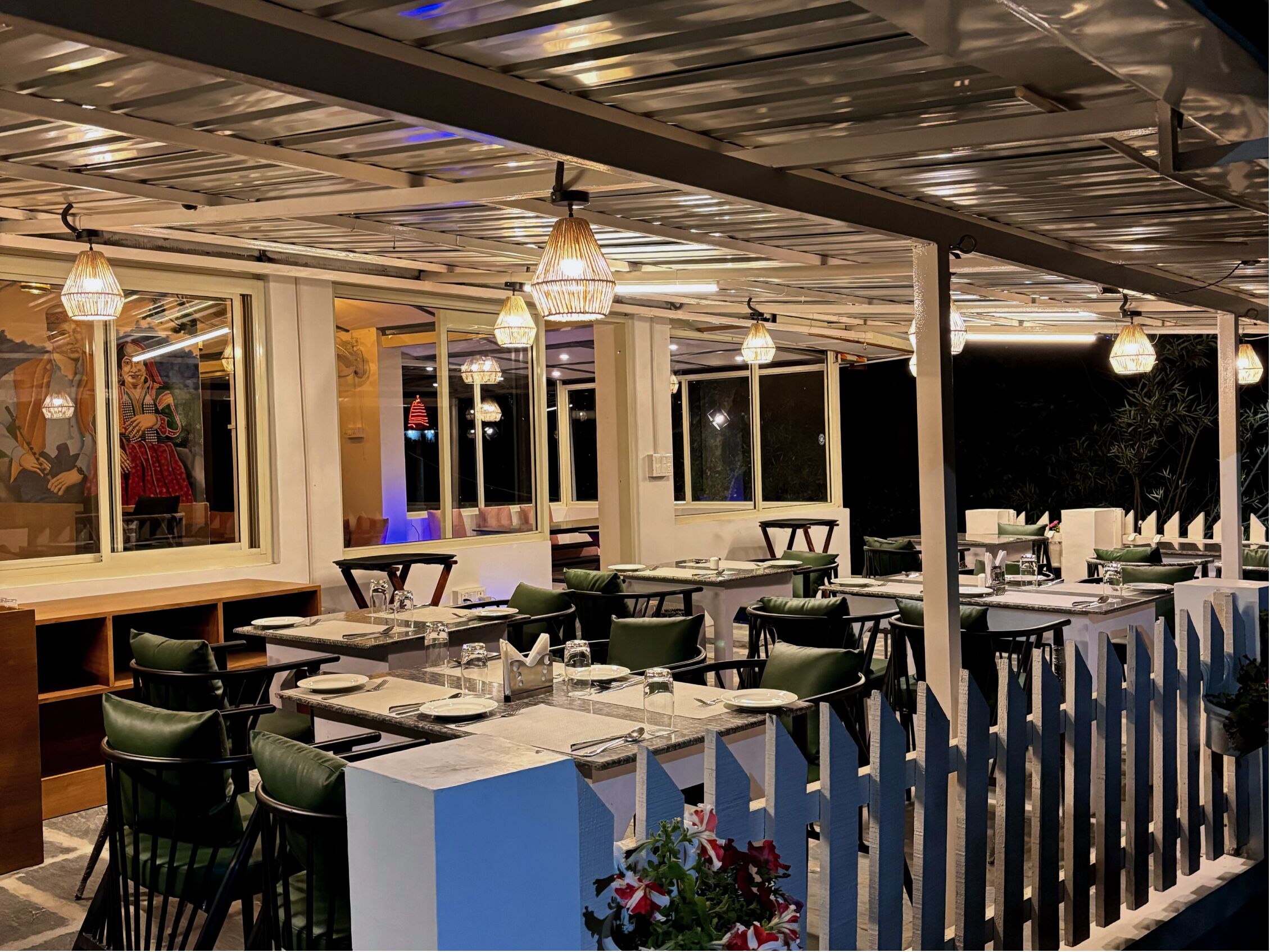 An outdoor restaurant with multiple tables and chairs, a counter with a menu, and hanging pendant lights.