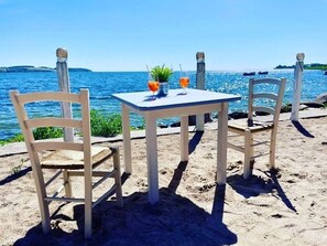 A beachside table with two chairs and a small table, overlooking a calm sea with boats in the distance.
