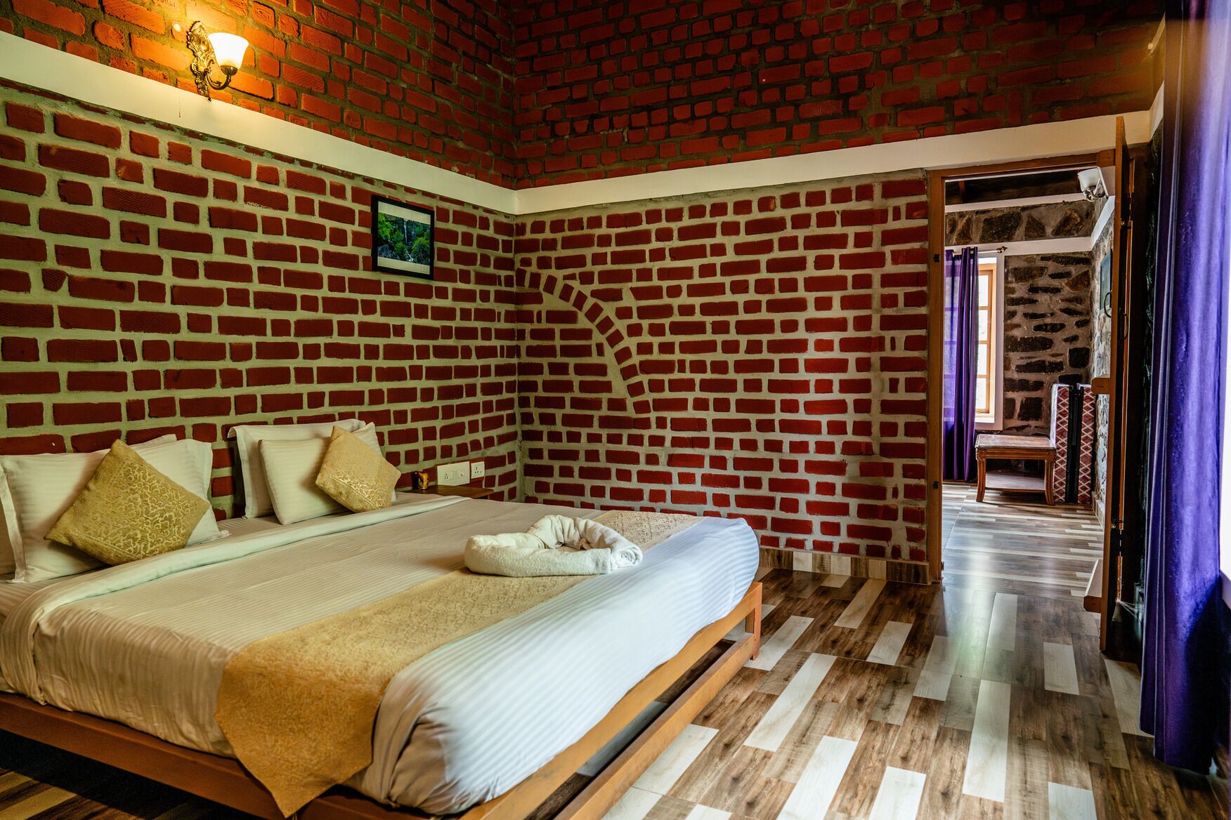 A bedroom with a brick wall, a bed with a white mattress and beige bedding, a wooden bench, and a stone wall in the background.