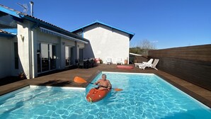 Une piscine où une personne fait du kayak, un patio couvert et un bâtiment avec un toit bleu.