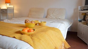 A bedroom with a bed, a bedside table with a lamp, a shelf with books, and a bowl of oranges on the bed.