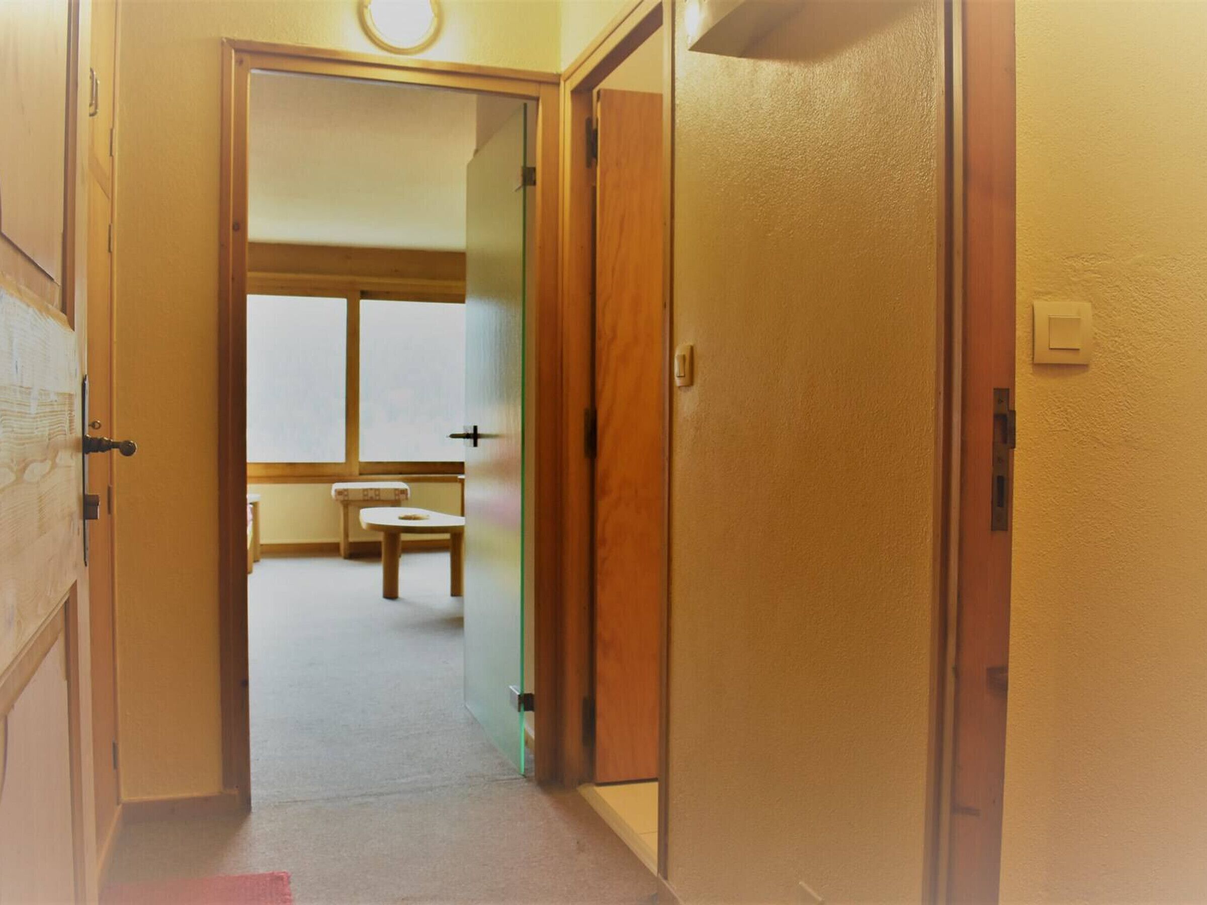 Un couloir menant à une pièce dotée d’une fenêtre, d’une petite table et d’une porte avec un interrupteur.