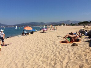Ein Strand mit Menschen, die sich sonnen und schwimmen, Sonnenschirmen und Segelbooten im Hintergrund.