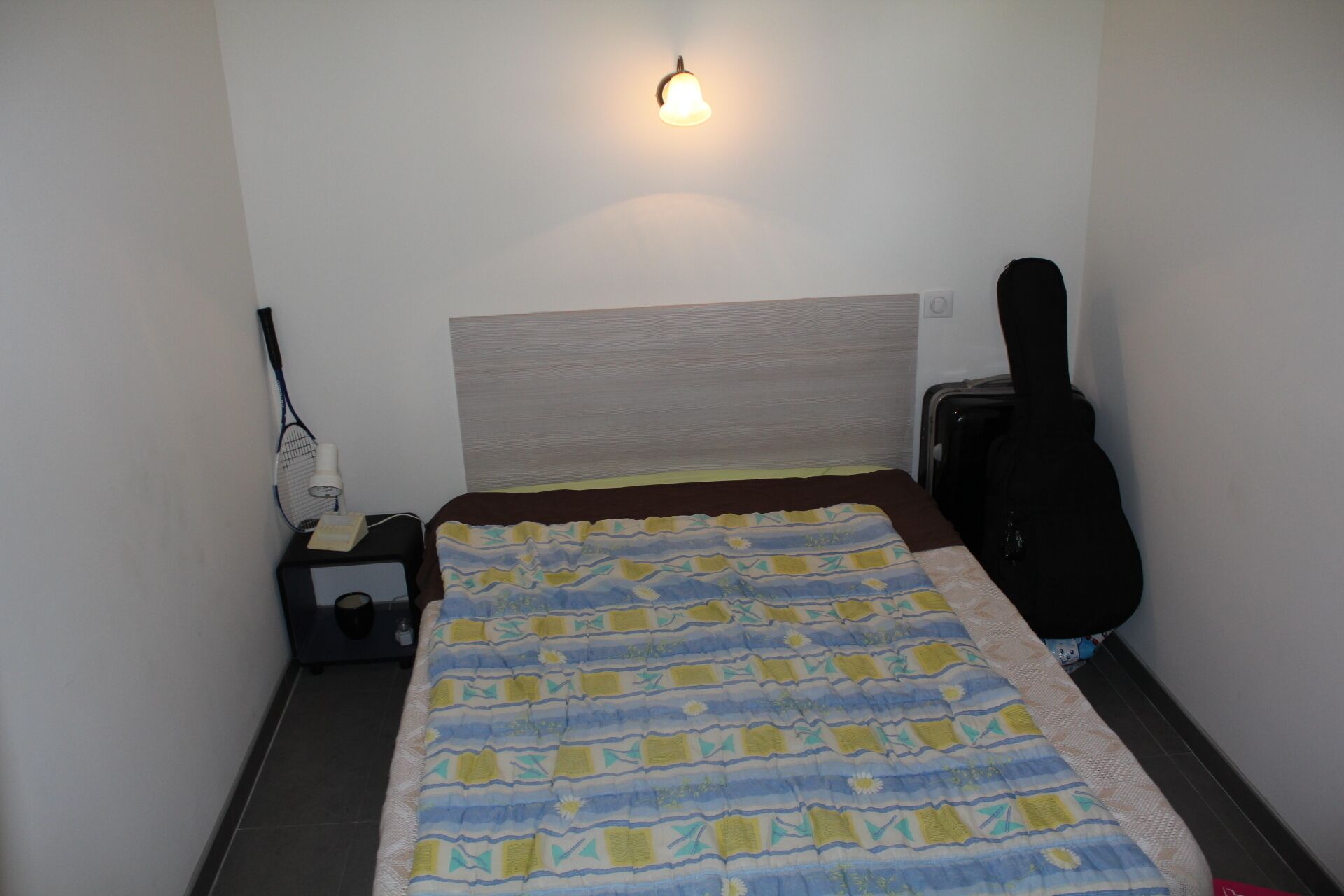 Ein kleines Hotelzimmer mit einem Bett, einer Gitarre, einer Lampe und einer Wandleuchte.