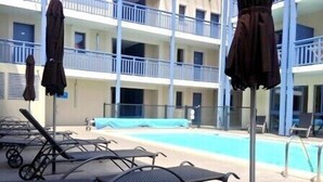Zona junto a la piscina con sillones de descanso y sombrillas, una piscina y un edificio de varios pisos con balcones.