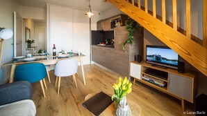 Una sala moderna con una escalera de madera, un comedor con una mesa dispuesta para cuatro personas, una cocina con horno empotrado y un televisor sobre un mueble de madera.