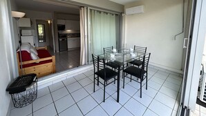 A compact living space with a glass dining table and chairs, a small kitchenette, and a sliding glass door leading to a bedroom with a red bedspread. - Air-Conditioned Studio with Terrace, Pool & Parking - Sleeps 4, Steps from Port Camargue Beach (Port Camargue)