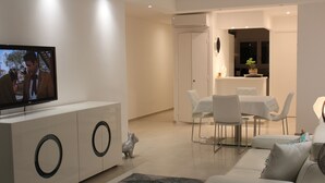 A modern living room with a flat-screen TV, a sofa, a dining table with chairs, and a kitchen area in the background.