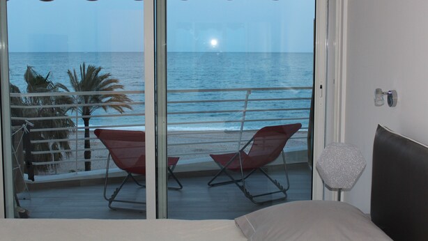 A room with a balcony overlooking the sea, featuring a bed, two red chairs, and a small table.