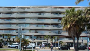 A multi-story building with balconies, palm trees, and a street in front.