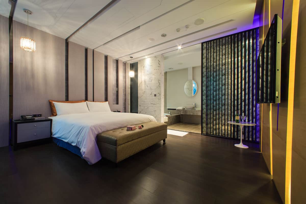 Un dormitorio moderno con una cama grande, mesitas de noche, un banco, un baño con espejo y un panel decorativo en la pared.