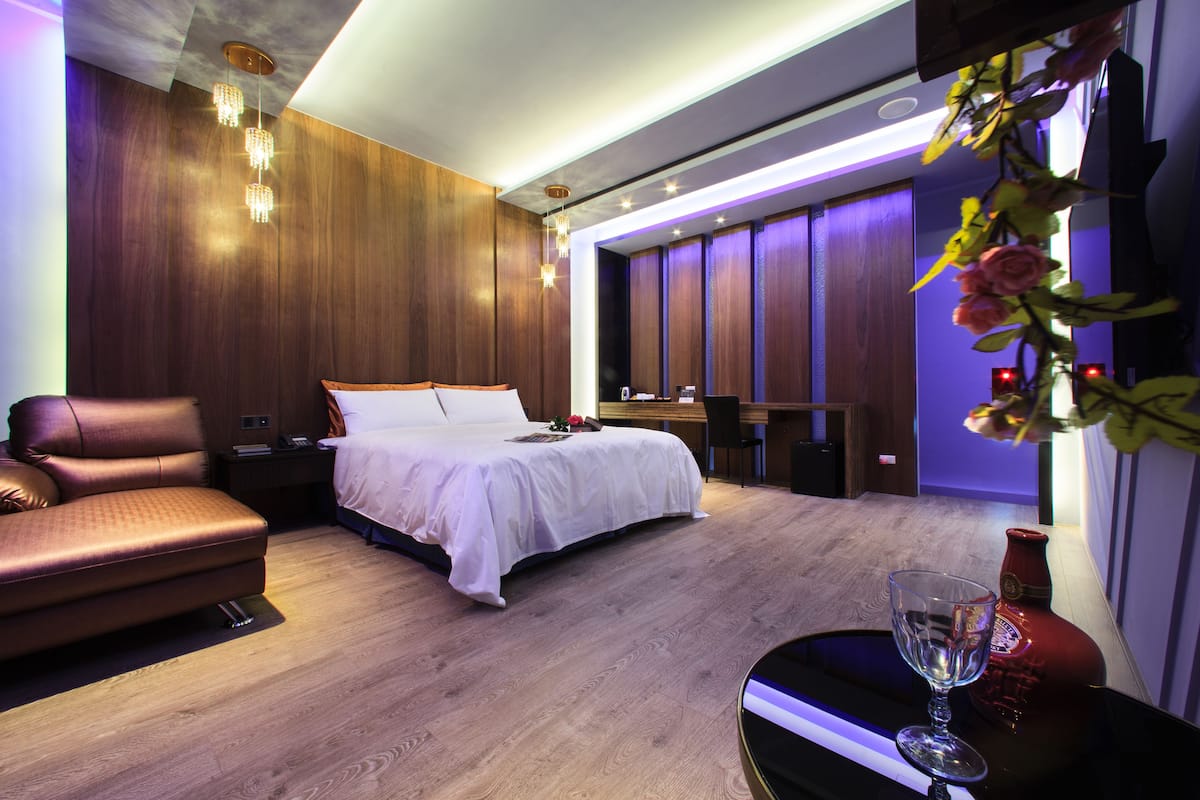 Una habitación de hotel moderna con suelo de madera, cama con ropa blanca, sofá de cuero, escritorio con silla y un jarrón decorativo.