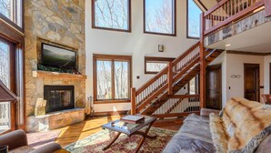 A spacious living room with a stone fireplace, a large window, a wooden staircase, and a leather sofa.