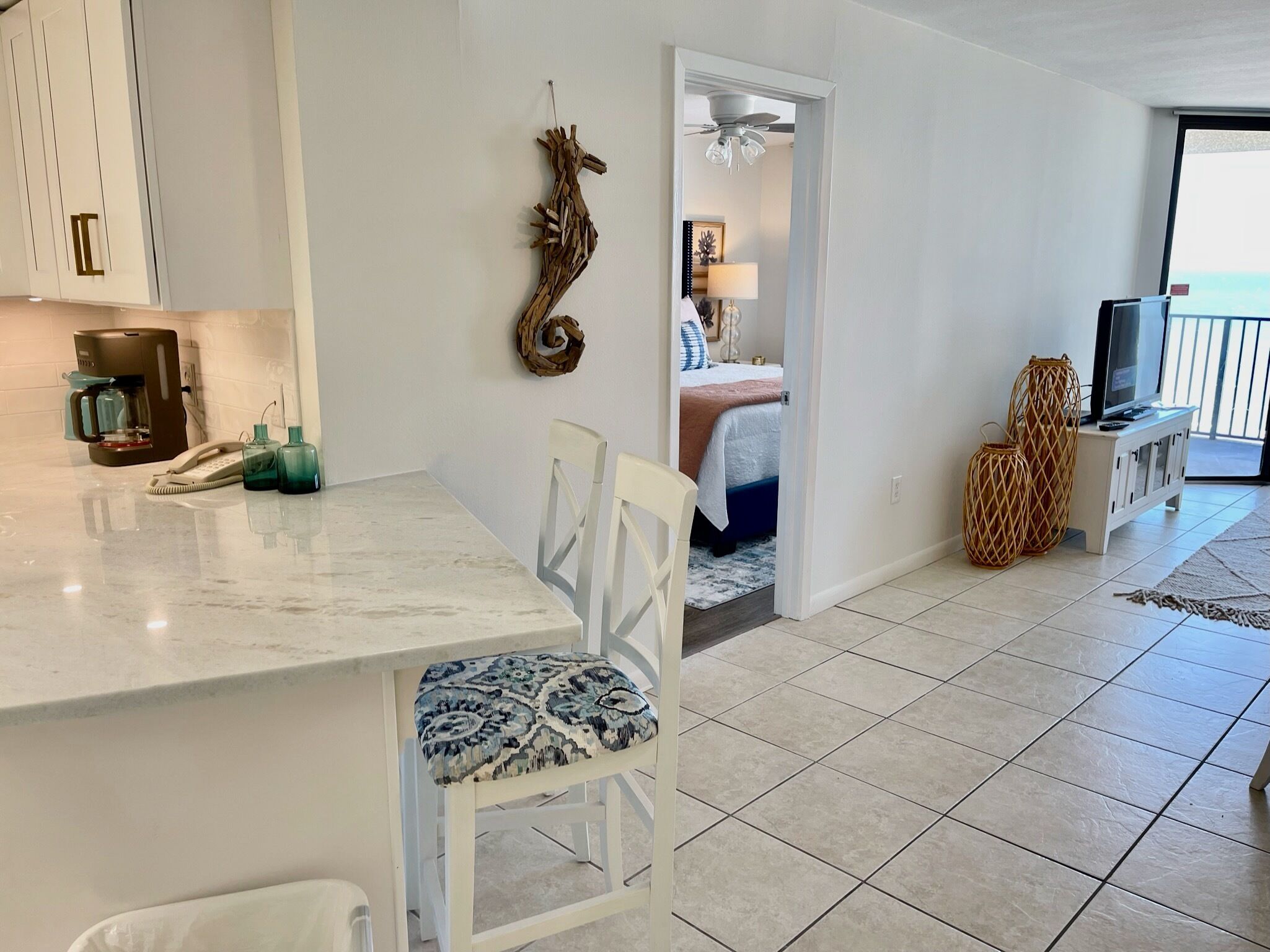 A modern kitchen with a marble countertop, a coffee machine, and a phone. There is a dining area with a patterned chair and a white table. A bedroom with a bed and a TV is visible through an open door.