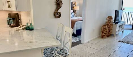 A modern kitchen with a marble countertop, a coffee machine, and a phone. There is a dining area with a patterned chair and a white table. A bedroom with a bed and a TV is visible through an open door.