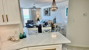 Una cocina moderna con muebles blancos y pomos dorados, encimera de mármol y vistas al área de estar, que incluye un sofá y una mesa de comedor.