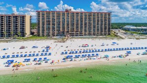 Un hotel frente a la playa con varios edificios altos, una playa arenosa con numerosas sombrillas y un cielo azul despejado.