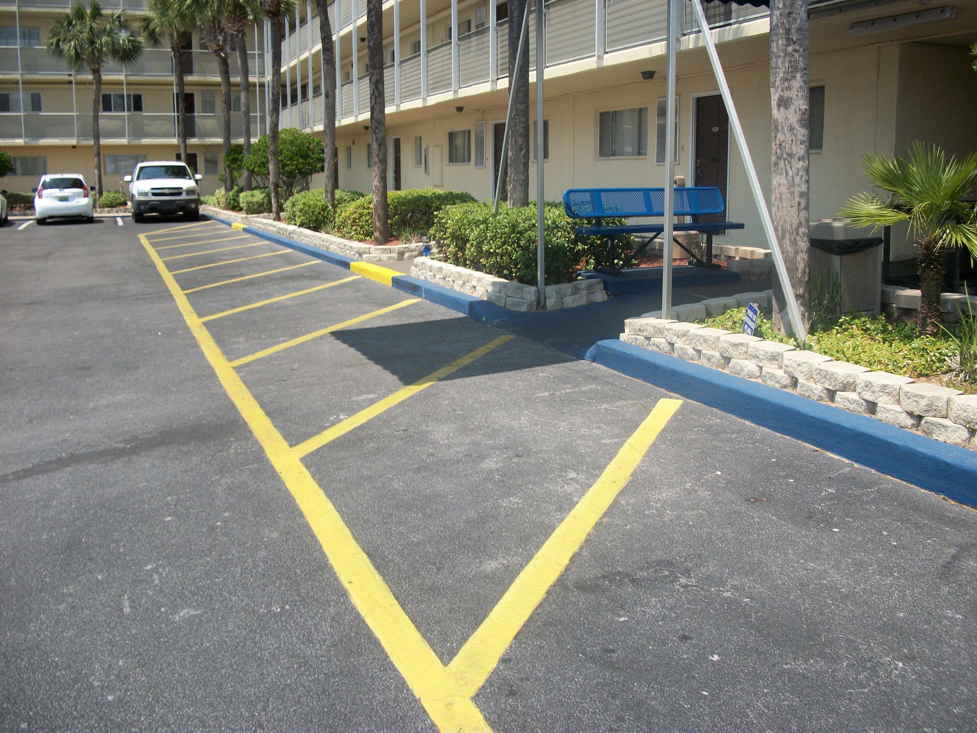 Un parking avec des places délimitées, un banc bleu et un bâtiment doté d’un balcon.