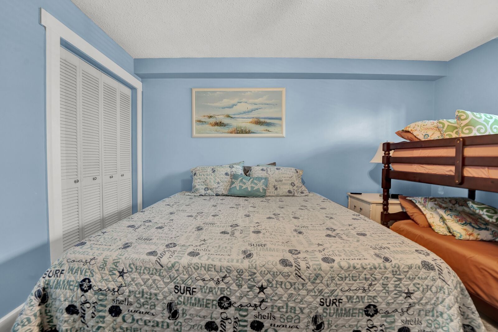 A bedroom with a bunk bed, a bed with a patterned bedspread, a painting on the wall, and a closet with louvered doors.