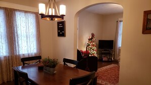 A dining area with a wooden table and chairs, a chandelier, a Christmas tree, and a television in the background.