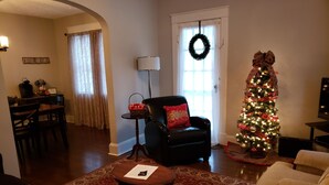 A living room with a Christmas tree, a black leather chair, a coffee table, and a dining area with a table and chairs.