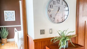 Un pasillo con piso de madera, un reloj de pared grande, un helecho en maceta y un cuadro en la pared.