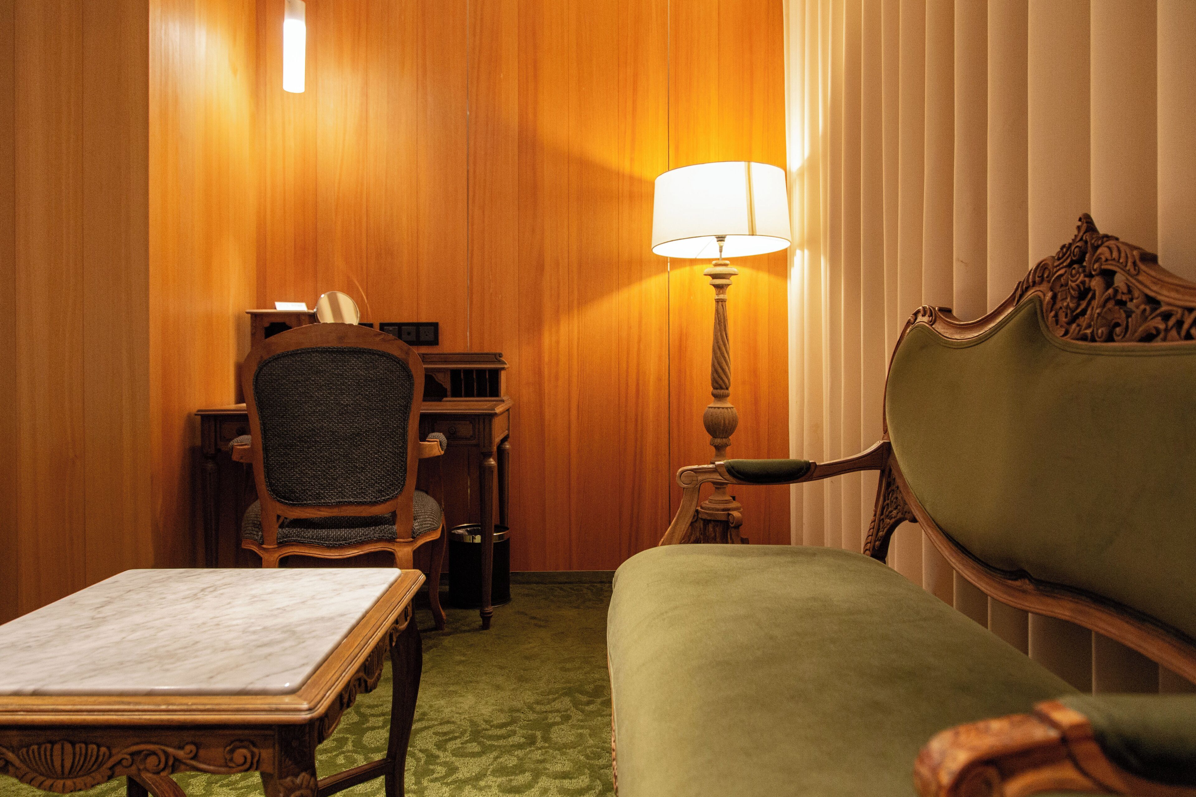 Una habitación con un revestimiento de madera, un sofá tapizado en verde, una mesa de madera con cubierta de mármol, una silla y una lámpara de pie.