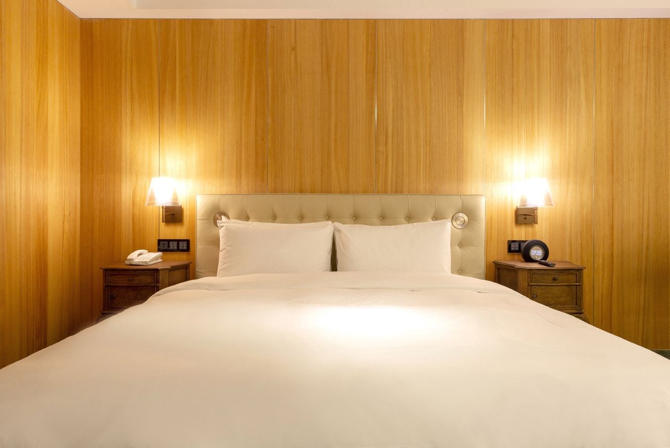 Habitación de hotel con una cama grande, paneles de madera en las paredes, dos mesitas de noche con lámparas y un reloj sobre una de ellas.